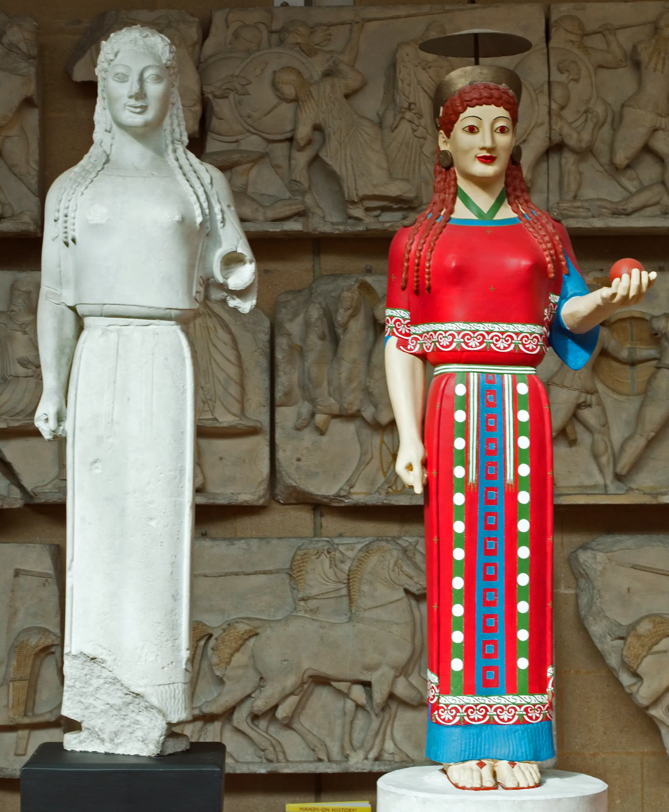 Zwei Museumsabgüsse der antiken griechischen Peplos-Kore nebeneinander: ein heller Gipsabguss und eine Rekonstruktion mit bemalten Haaren, Haut, Kleidung und gemusterter Verzierung.